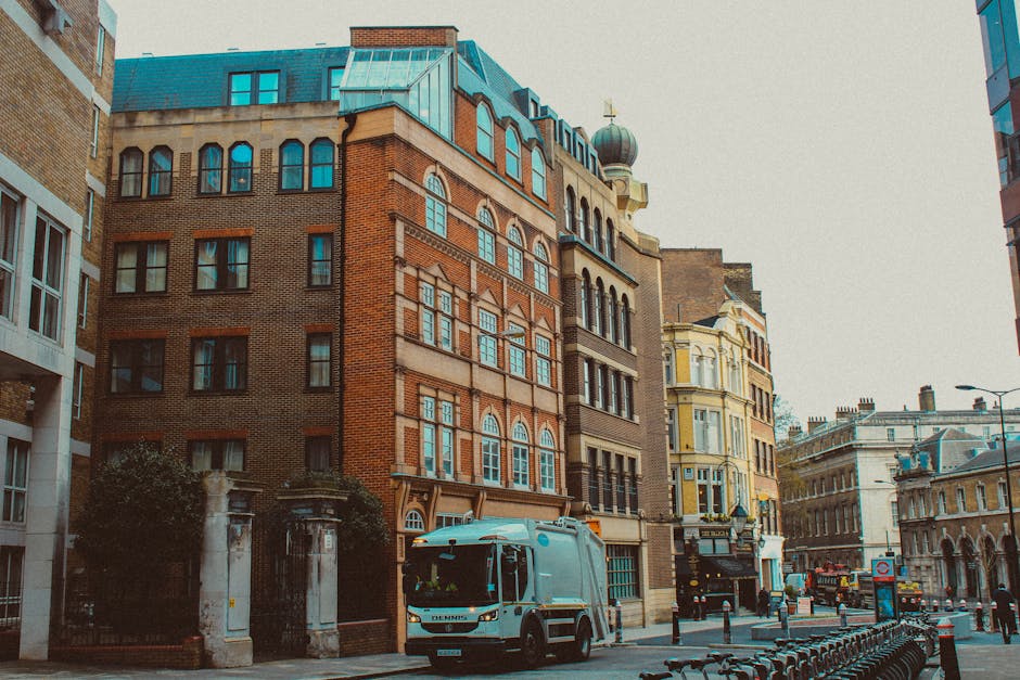 A large moving truck with a green cargo area is parked on a city street in front of tall modern skyscrapers and older stone buildings, indicating an urban area suitable for home relocation or furnitur