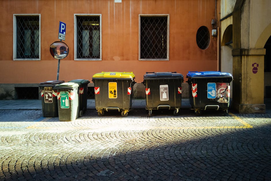 A person wearing a light grey T-shirt and beige cap is standing beside a small green waste collection truck on a street during daylight. The individual is reaching up with their right hand, possibly t