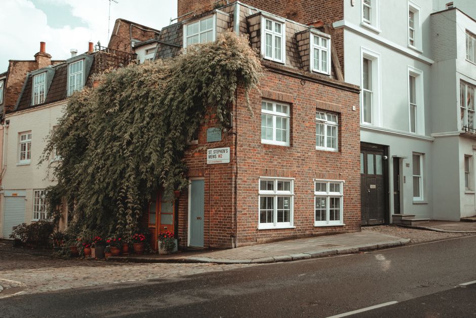 A view through a brick archway labeled 'Hyde Park Gardens Mews W2' leading into a narrow cobbled street lined with white and beige terraced houses with small front gardens and potted plants. The stree