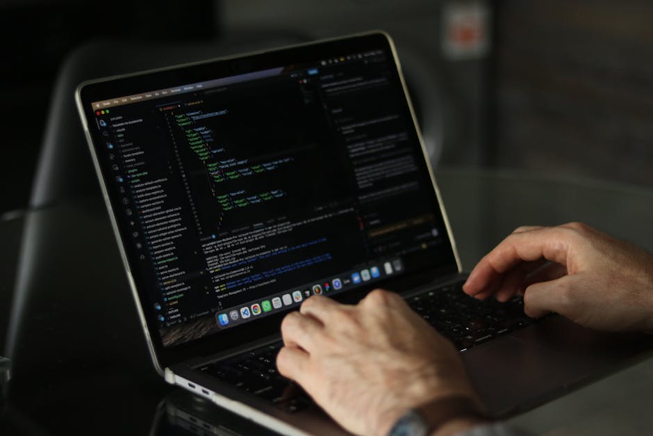 A person's hand, wearing a wristwatch, is positioned on a keyboard of a silver laptop placed on a wooden surface. The laptop screen displays lines of HTML code, indicating activity related to web deve